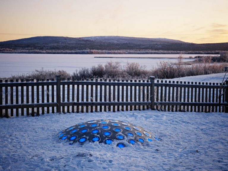 Termonodens lampor lyser blå i den snötäckta gryningen. Bakom staketet sträcker sig fjället. Markus Lantto, Termonoder, 2009