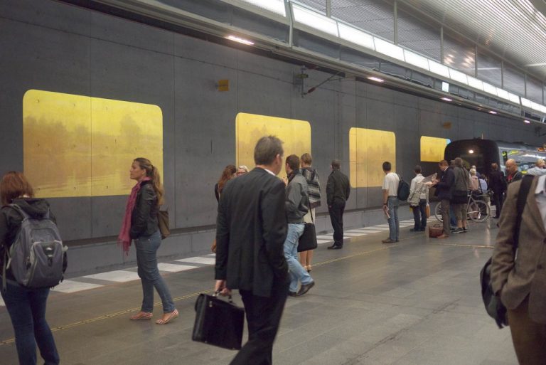 Människor väntar på tåget som kör in vid perrongen. På väggen projektioner av vatten och träd i dis. Tania Ruiz Gutiérrez, Annorstädes