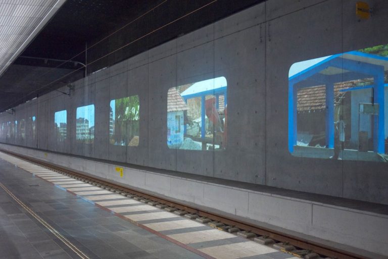 På betongväggen bakom en tågräls syns flera projektioner av bilder med hus på. Tania Ruiz Gutiérrez, Annorstädes