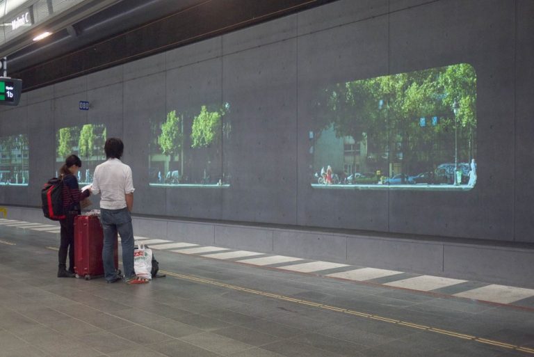 Två personer med packning väntar på perrongen och tittar på projektioner av stadsmiljöer. Tania Ruiz Gutiérrez, Annorstädes