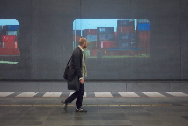 En man går på perrongen och tittar på en projektion av färgade containrar som ligger staplade på varandra. Tania Ruiz Gutiérrez, Annorstädes