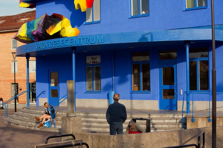 Detalj av blåmålade Vara Resecentrum, med färgglada objekt på taket. Människor som tittar. Katharina Grosse, Blue Orange, 2012
