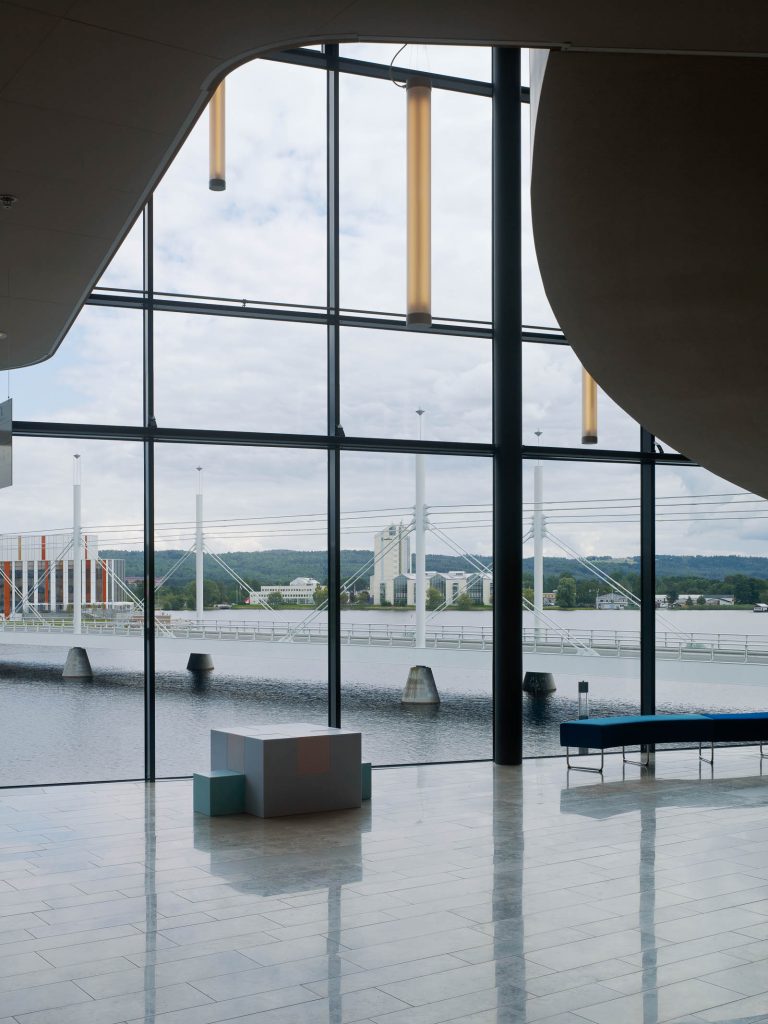 Inifrån byggnaden genom glasfasaden syns vattnet och en lång järnbro. Innanför fönstret en vit och blå skulptur. Jacob Dahlgren, Tetris, 2012.