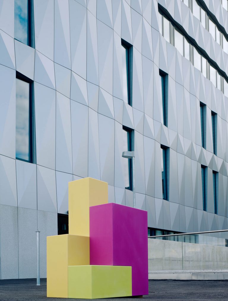 Skulptur utomhus med rosa och gula rätblock på högkant och ett litet grönt block vid foten. Jacob Dahlgren, Tetris, 2012.