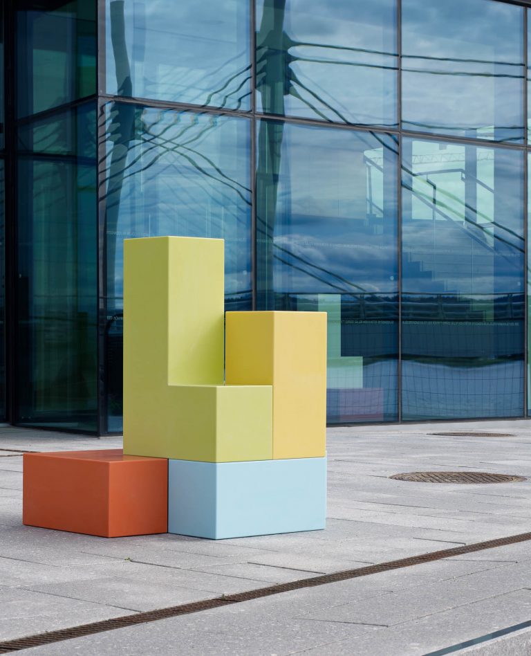 Skulptur utomhus. Ett blått och ett orange block i basen och ett gult och ett grönt block placerade ovanpå. Jacob Dahlgren, Tetris, 2012.