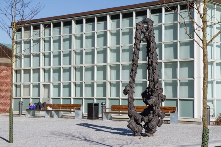 Hög bronsskulptur. I bakgrunden bänkar och en byggnad. Carl Boutard, Into The Wild (2013)