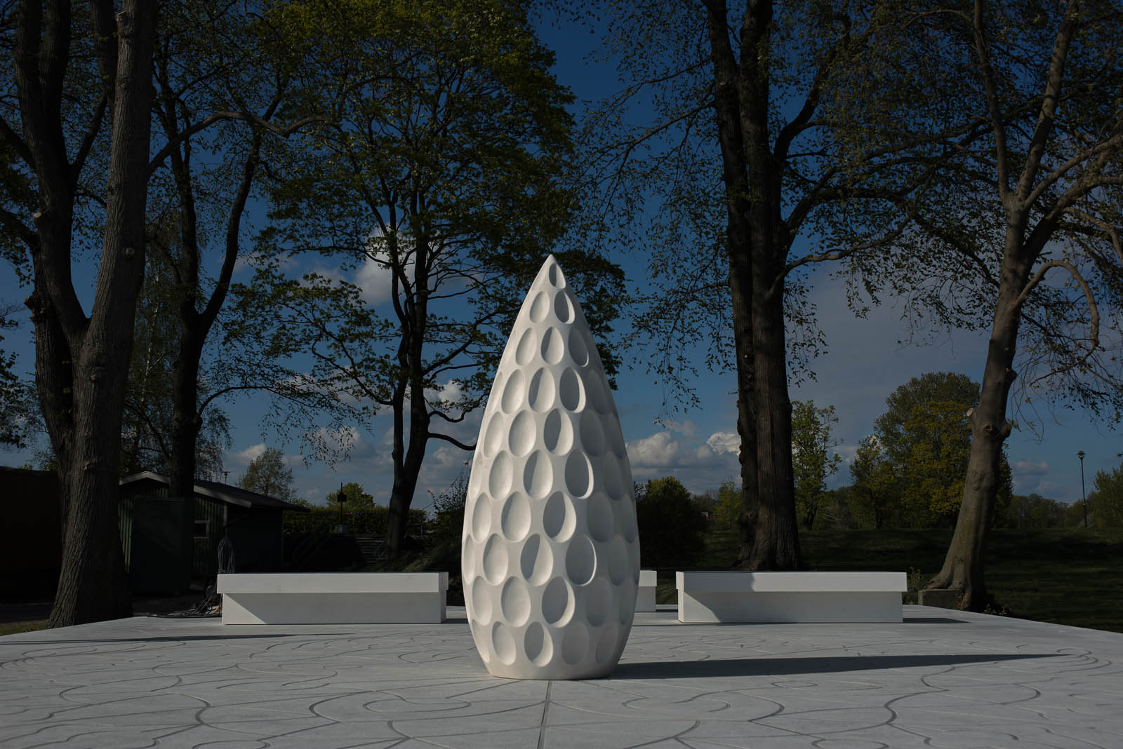 En hög vit stenskulptur, med formen av ett utdraget ägg. Skulpturen står på ett vitt stengolv, med vita bänkar bredvid sig. Monika Larsen Dennis, Restare, 2013