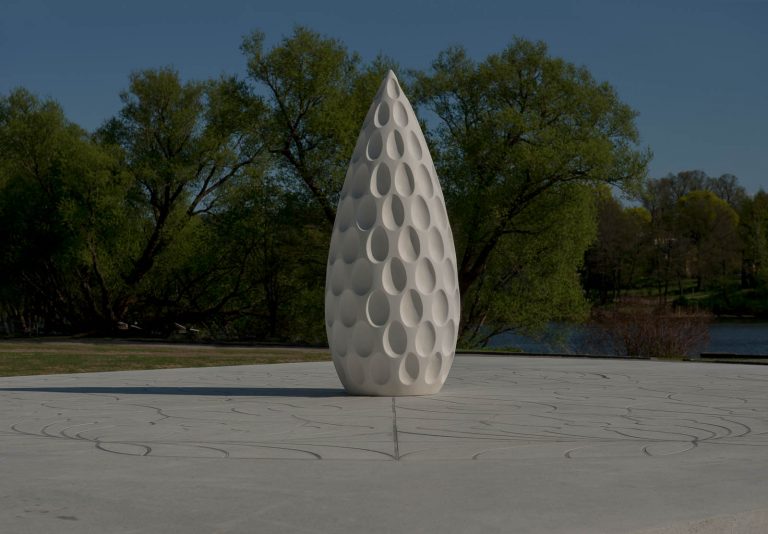 Den vita skulpturen är mönstrad med ovala urholkningar. Även det vita stenunderlaget har ett slingrande mönster. I bakgrunden träd och vatten. Monika Larsen Dennis, Restare, 2013