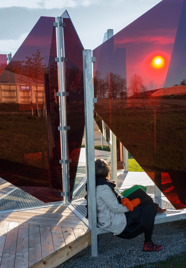 En kvinna sitter bortvänd på trägolvet mellan glasskivorna som speglar den sjunkande solen. Patrik Aarnivaara, Tidsglänta, 2013.