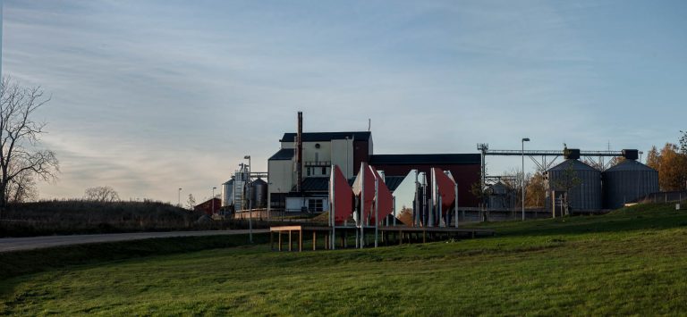 Verket står på en gräsmatta. I bakgrunden biogasanläggningen med skorsten och grå cisterner. Patrik Aarnivaara, Tidsglänta, 2013.