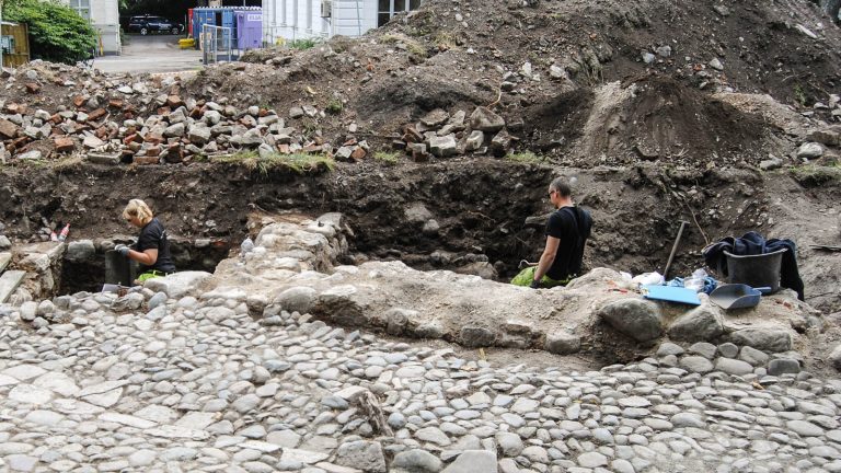 En kvinna och en man i arbetskläder står upp till midjan i varsitt hål och arbetar med utgrävningen. Vid sidan jordmassor och uppkastade stenar. Kvarteret Valnötsträdet, utgrävningar 2013