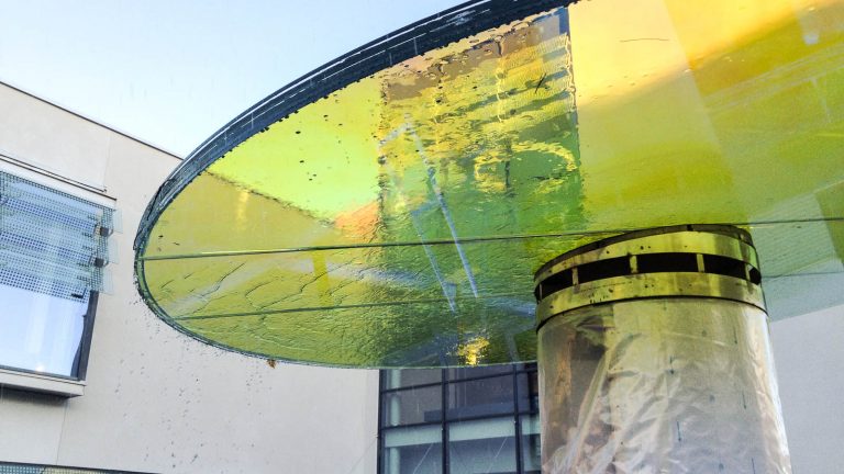 Det runda glaset över fontänen och seglen är laminerat med en folie som reflekterar, speglar och bryter ljus. Fontänens/källans glastak ser gult ut på bilden. Torbjörn Johansson, Källan Är