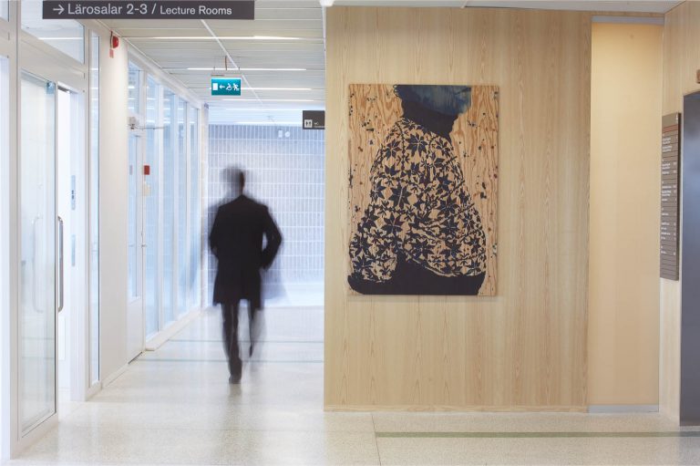 En person rör sig genom en korridor. På en trävägg hänger en tavla med en korpulent överkropp i mönstrad polokofta, målad på trä. Robert Lucander, 13 målningar