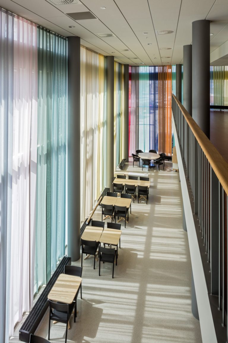 Korridor med stolar och bord. Ena väggen är täckt från golv till tak av ett flerfärgat draperi som släpper igenom ljuset utifrån. Astrid Krogh, Layers of Colour and Light
