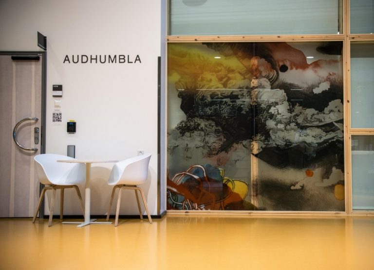 Två stolar och ett bord i en korridor bredvid en stor målning på en glasvägg, med färgade formationer. Christoffer Paues, All Världens Djur