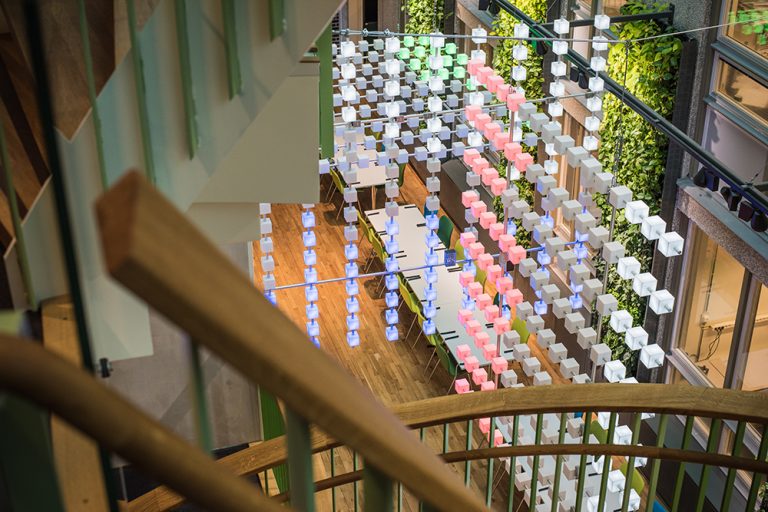 I ljusgården nedanför ett trappräcke hänger serier med kuber i olika färger monterade i lodräta och vertikala rader. Gustav Hellberg, XYZ
