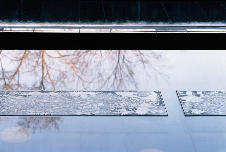 Stavar av isrosat glas placerade i en grund vattenspegel. I vattnet speglas trädgrenar och himmel. Ingegerd Råman, March 6 a.m.