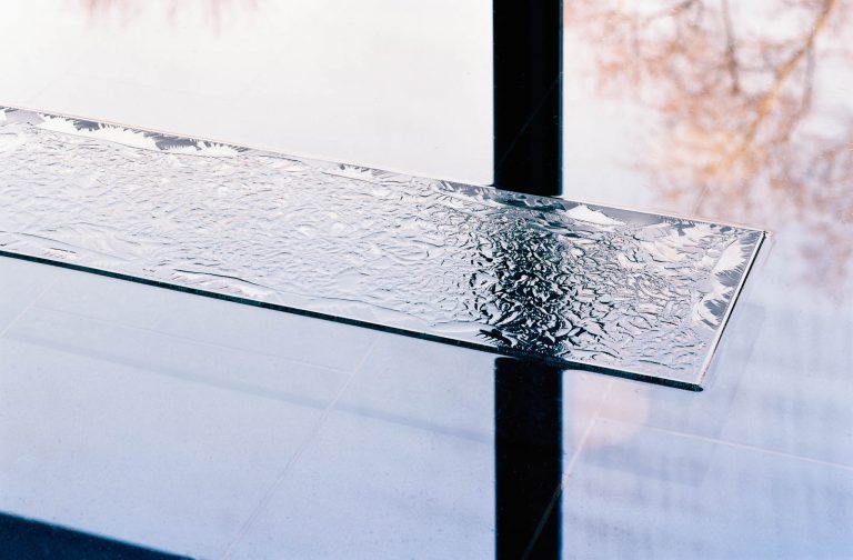 Stavar av isrosat glas i en grund vattenspegel på svart granit. I vattnet speglas trappräcket till nästa våning och de höga ljusa fönstren. Ingegerd Råman, March 6 a.m.