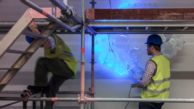 En man i reflexväst testar den turkosskimrande belysningen från en glasformation på väggen. Fredrika Linder, W