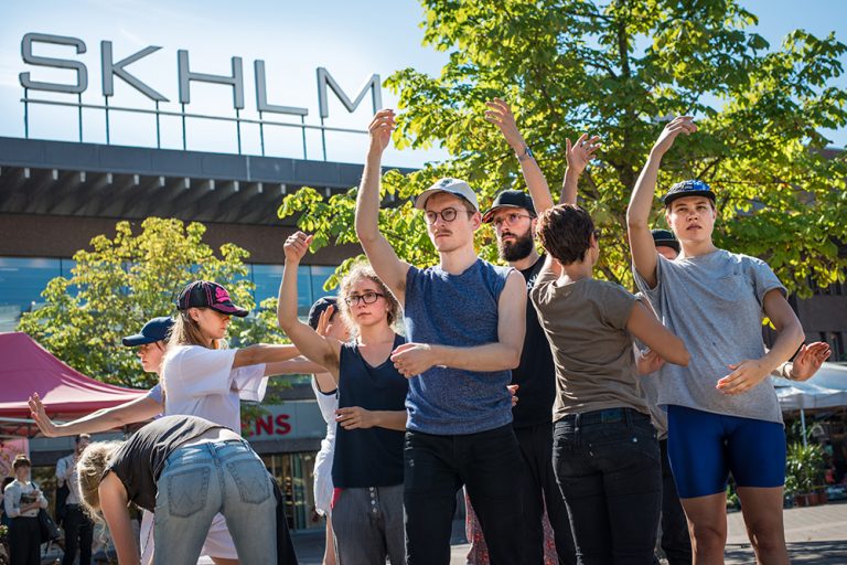 En grupp personer står utomhus vända åt olika håll med ena armen lyftad. Arbetets monument Alexandra Pirici