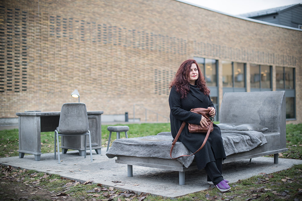 En kvinna med rött hår sitter på en betongsäng som står på en betongplatta på gräsmattan framför ett tegelhus. Mandana Moghaddam, Vinden bär oss med sig