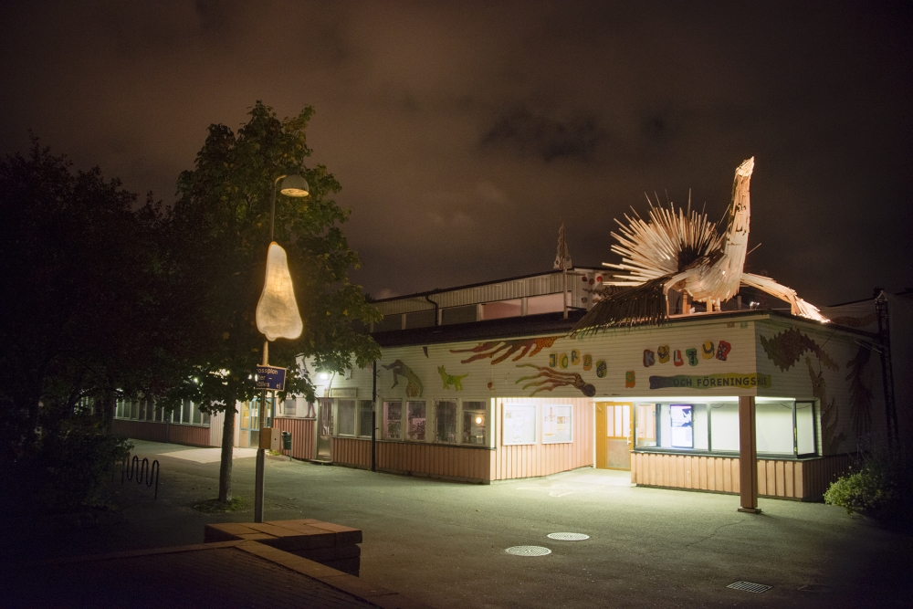 En lysande näsa på en lyktstolpe till vänster i bild och en belyst fågel på taket till kulturhuset i Jordbro. Det är kväll..