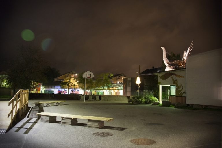 En sen höstkväll i Jordbro med vy över basketplanen utanför kulturhuset. Bakgrunden syns den lysande näsan och på husets tak tornar fågeln upp sig.