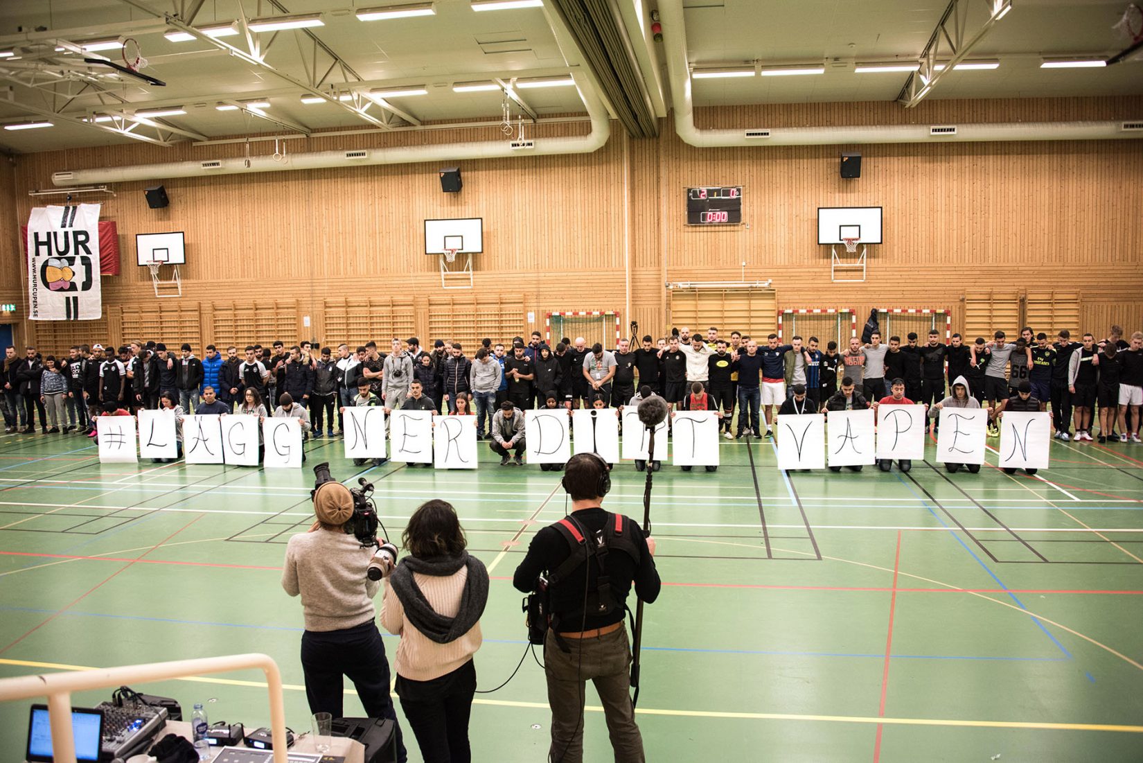 Ett stort antal unga män håller varandra om axlarna. Framför dem hukar andra ungdomar och håller upp texten "Lägg ner ditt vapen". Filmteamet dokumenterar.