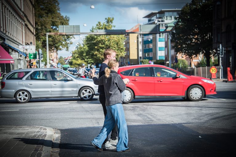 En kvinna leder en blundande man över vägen. I bakgrunden kör bilar. Myriam Lefkowitz, Walk, Hands, Eyes (Malmö)