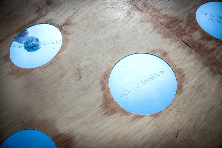 Detalj av ett trägolv, med två cirkulära speglar. I den ena ser man speglingen av en person. Där står texten: 