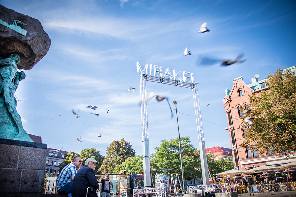 Ordet "Mirakel" i ljusskrift upphöjt på en ställning. I bakgrunden duvor och delar av Möllevångstorget. Santiago Mostyn, Mirakel.