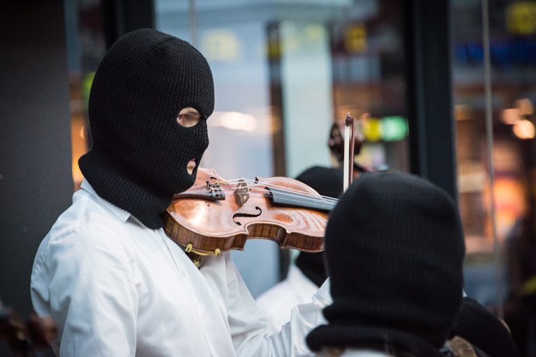 En violinist i svart heltäckande ansiktsmask, med hål endast för ögon och mun, står med fiolen på axeln och stråken lyft. Sislej Xhafa, Again and again.