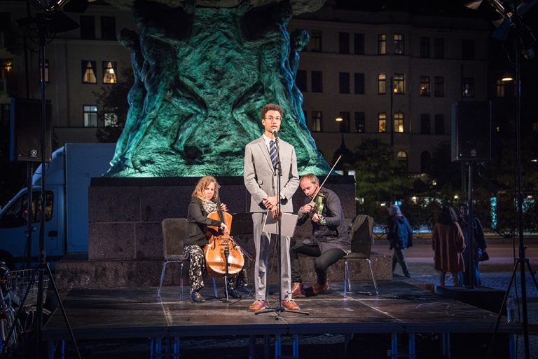 Konstnären Santiago Mostyn, iklädd slips och kostym, står på ett podium framför den upplysta statyn på Möllevångstorget. Bakom honom en cellist och en violinist. Santiago Mostyn, The repetition.