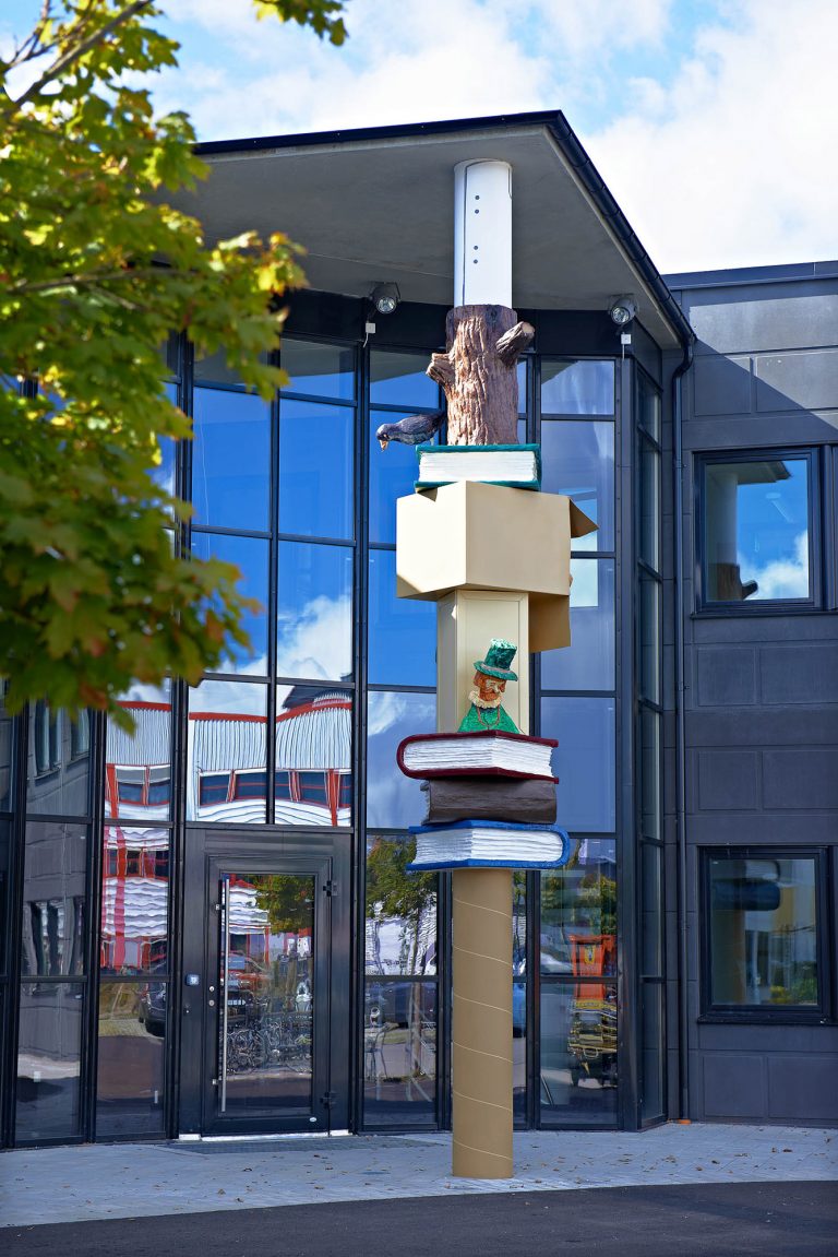 Pelaren med färgglada figurer står framför en hög glasfasad. Daniel Jensen, Fundamentet (2012)