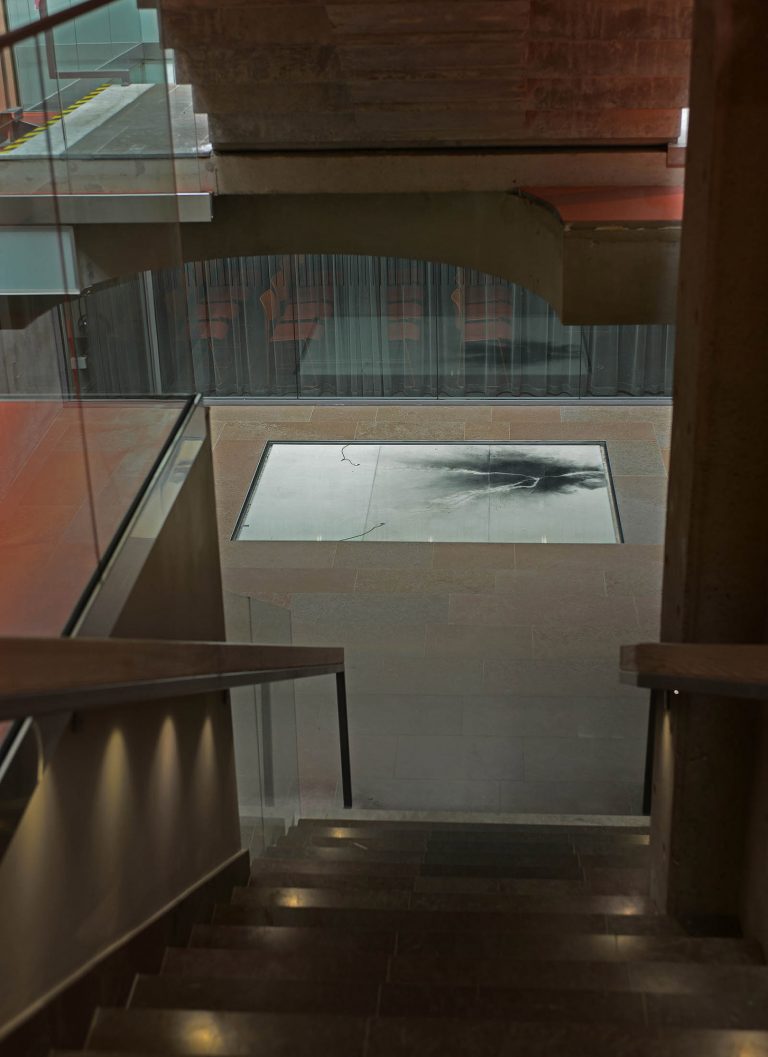 En trappa leder ner till den vita golvplattan med kol och två elsladdar. Nina Canell, Impulse Slight (100 000 Volt)