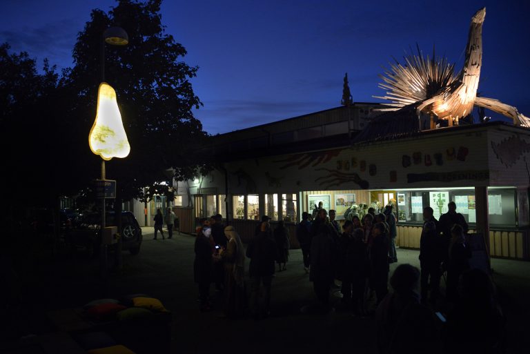 Människor i skymningen utanför kulturhuset i Jordbro, näsan på lyktstolpen är tänd och syns till vänster i bild, träskulpturen i form av en fågel står på kulturhusets tak till höger i bild.