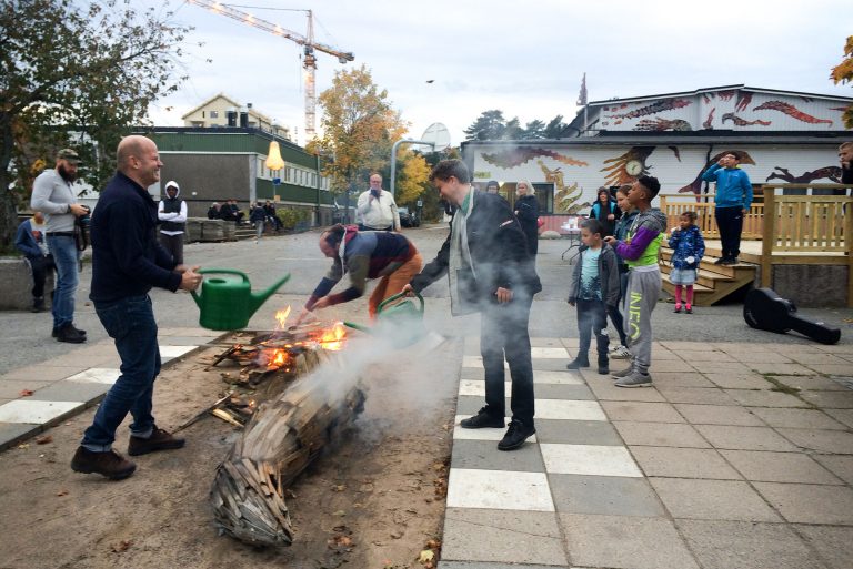 Människor står samlade framför en stor träskulptur i delar, vissa delar brinner samtidigt som två personer häller vatten på med gröna vattenkannor.