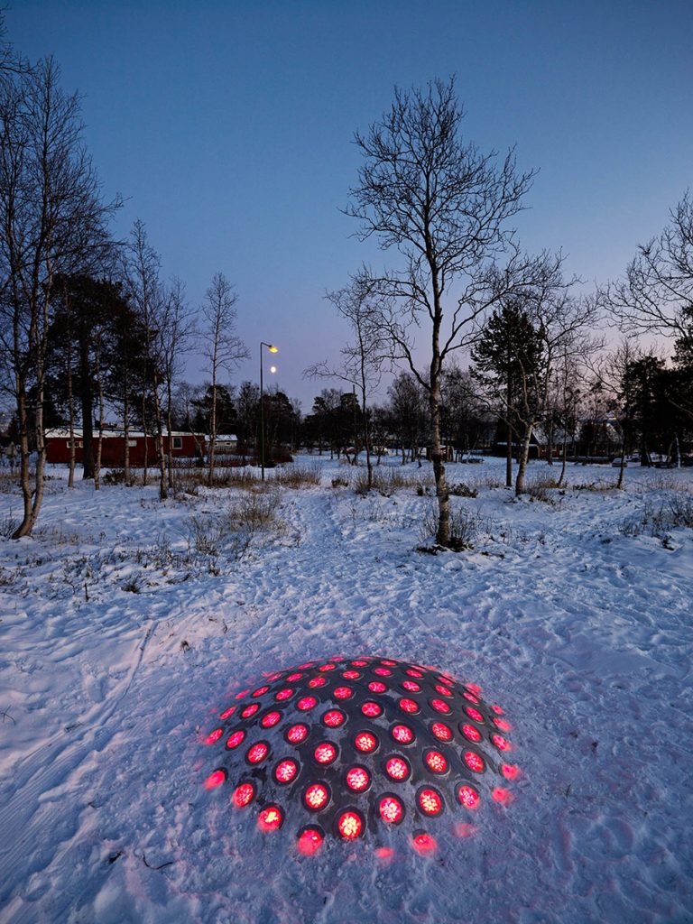 Ur den nedtrampade snön sticker termonoden upp. Lamporna lyser röda i mörkret. I bakgrunden träd och röda byggnader. Markus Lantto, Termonoder, 2009