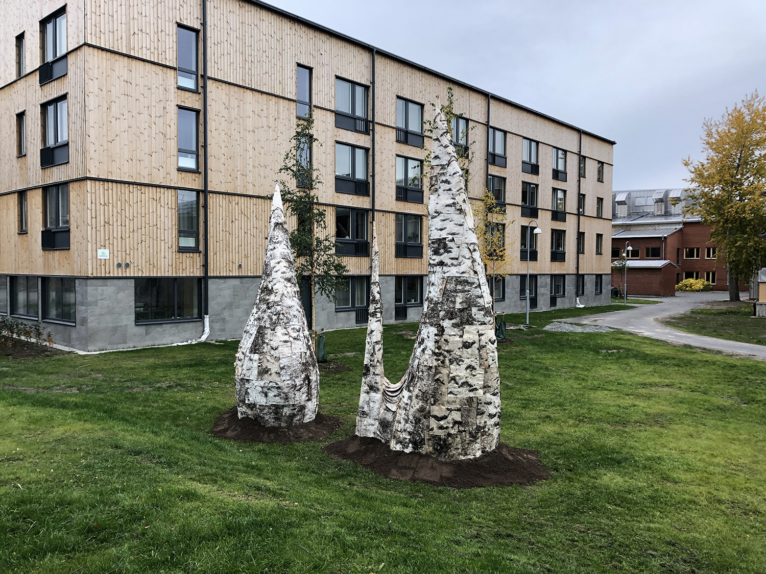 Sculptures made out of birch bark on a green lawn. There's a low building in the background.