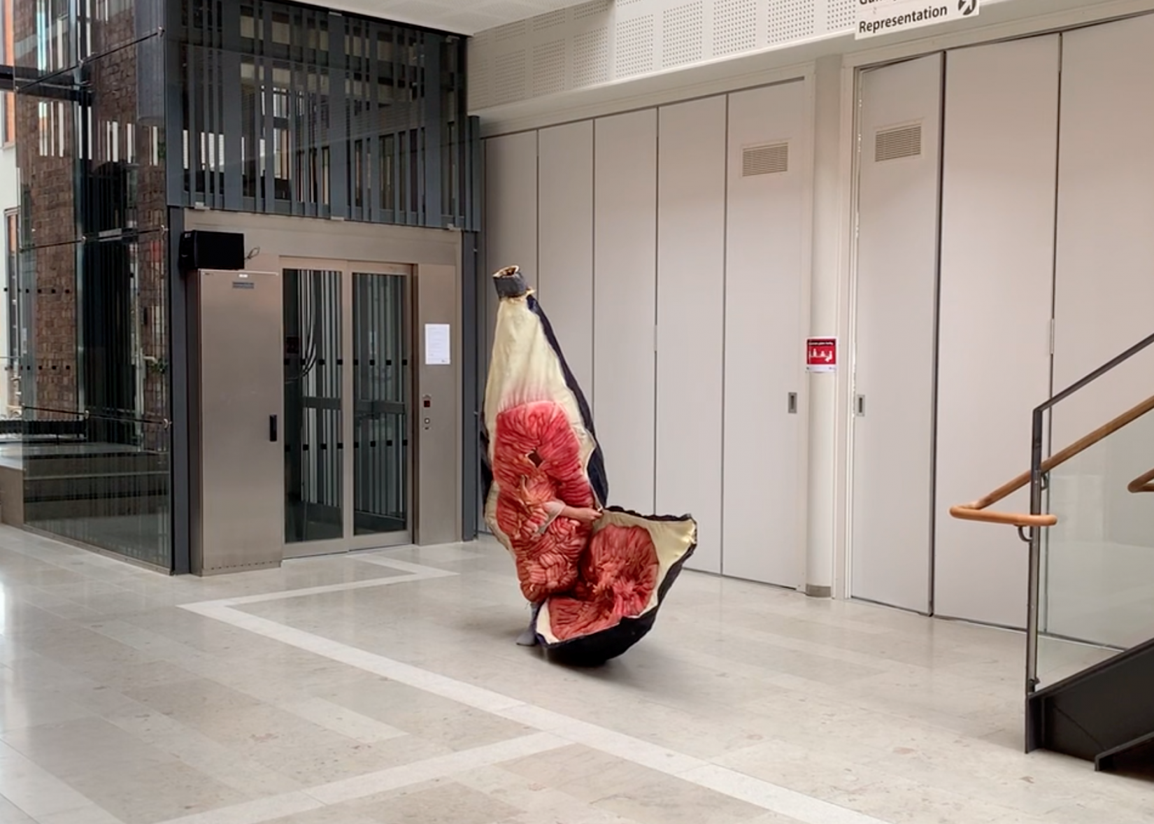 Mitt i en entrehall, framför dörrar och en hiss, syns ett skrynklig fikonliknande objekt. En arm sticker ut ur fikonets mitt.