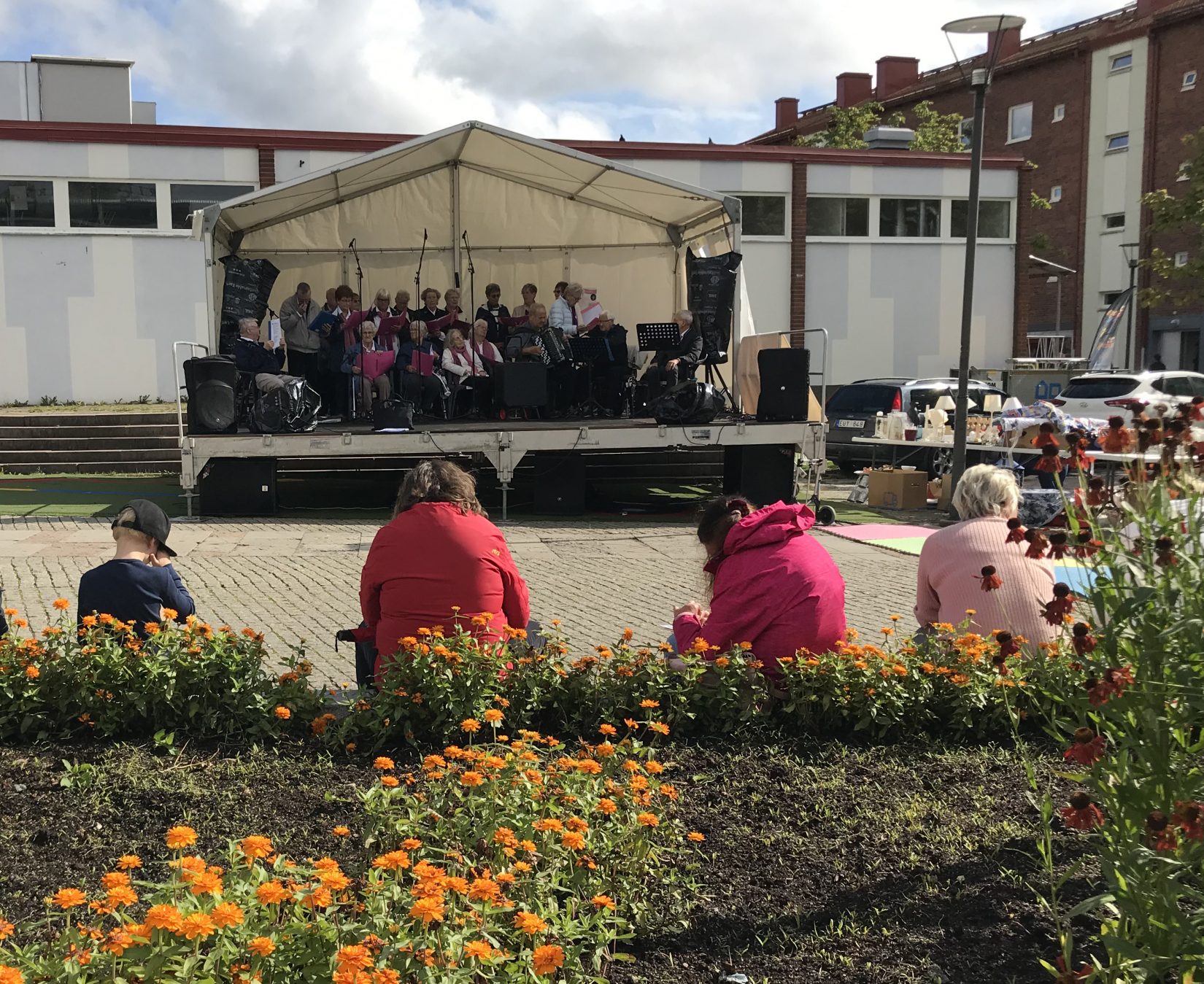 Fyra personer sitter framför en rabatt och tittar mot en scen. På scenen spelar en orkester.
