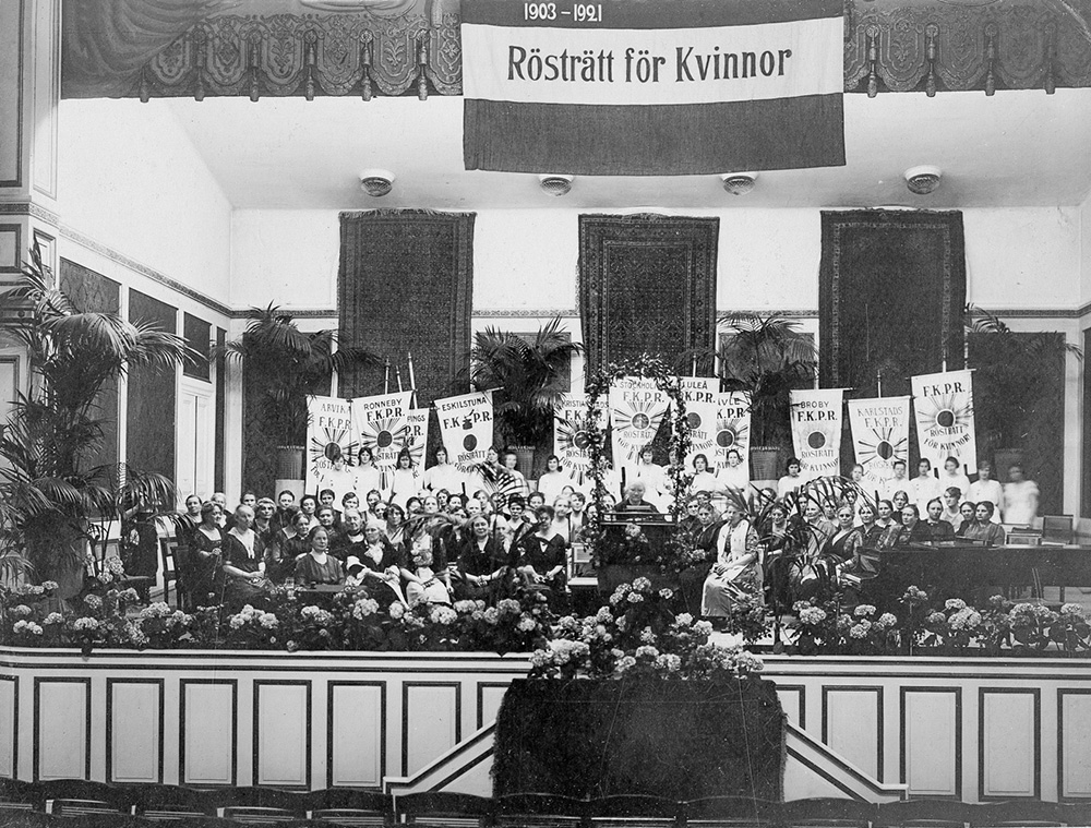 En svartvit bild på en scen. På scenen finns flaggor i bakgrunden och sittande kvinnor längre fram. En kvinna står vid en talarstor. Ovan scenen finns en flagga där det står Rösträtt för kvinnor.