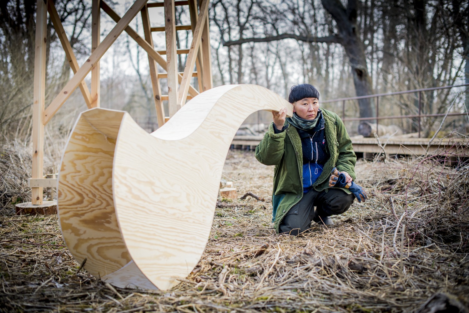 Konstnären sitter på huk och håller en stor lur i plywood mot örat.