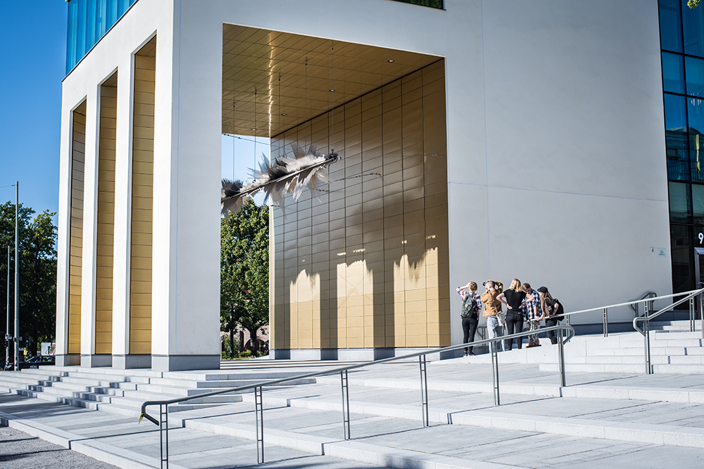 Skulptur som hänger i en guldfärgad loggia fotograferas av en grupp intresserade. Ebba Matz, Sch, tyst, lyssna