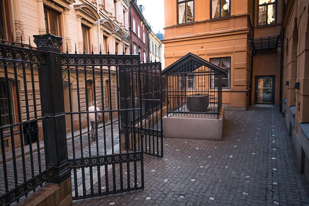 Öppna gallergrindar mot en stenlagd gård. En övertäckt träbåt omgärdad av metallgaller och med metalltak ovanför. Jan Håfström Arken