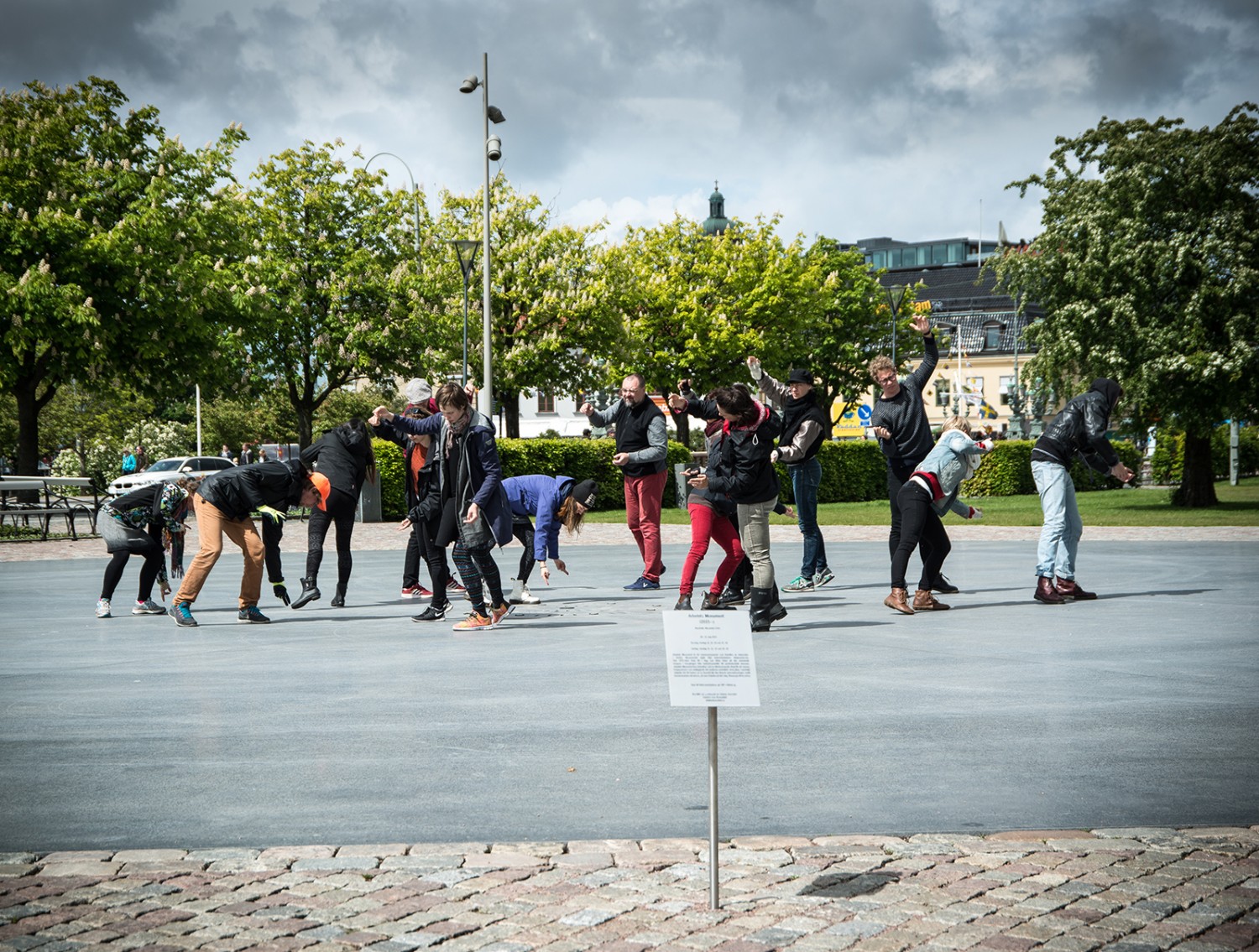 En grupp personer på en öppen plats. Alla står böjda åt olika håll och tycks vara i rörelse. Arbetets monument Alexandra Pirici