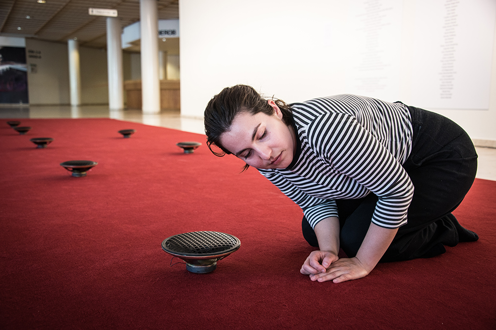 En kvinna sitter hopkrupen på en röd matta och lyssnar på ljudet från en av flera högtalare som finns utplacerade. James Webb, Prayer (Malmö)
