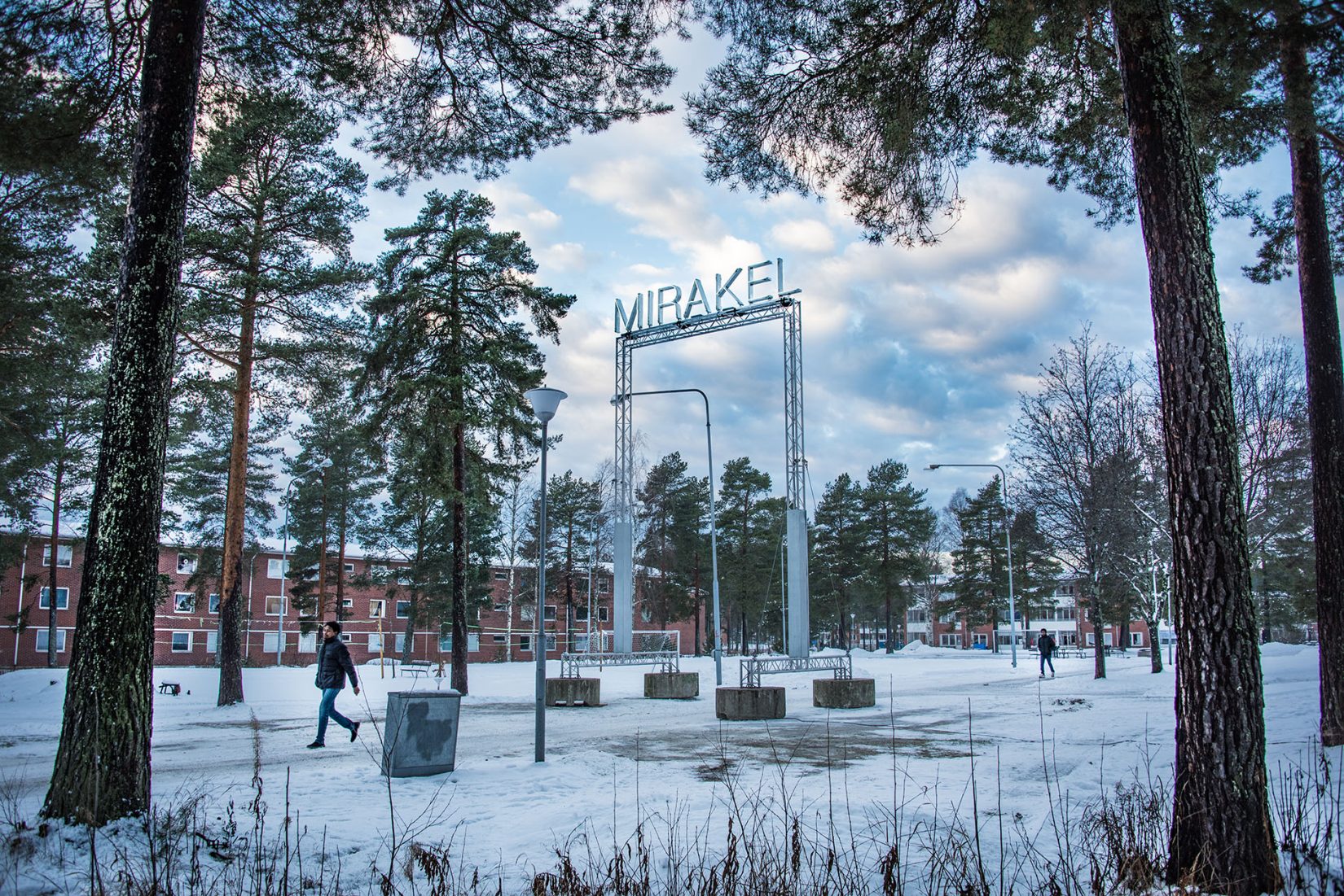 Ordet "mirakel" i ljusskrift högt uppe på en ställning i en skogsdunge. Bostadshus i bakgrunden. Santiago Mostyn, Mirakel