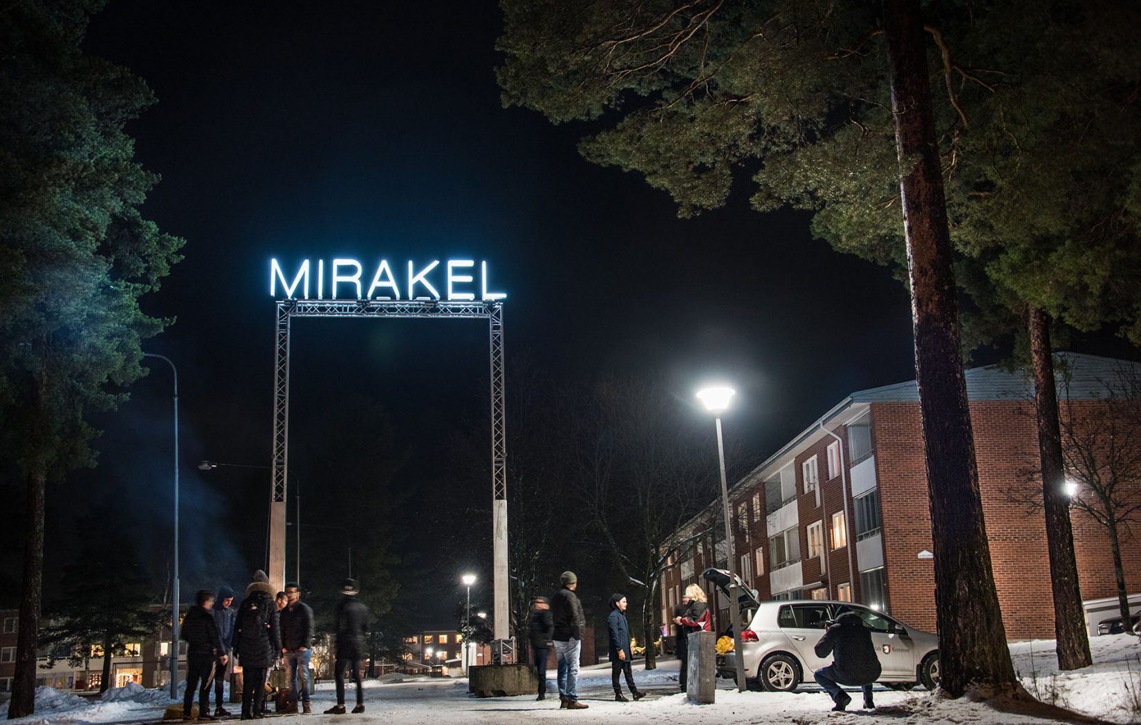 Ordet "mirakel" lyser mot en mörk natthimmel. Människor och ett bostadshus. Santiago Mostyn, Mirakel