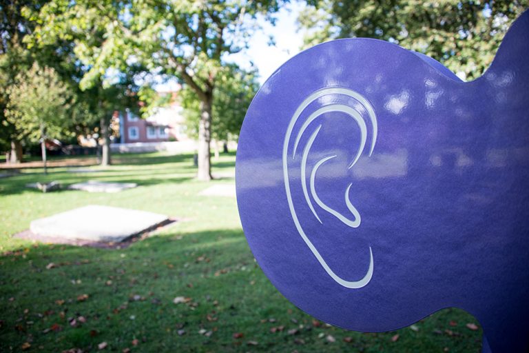 Ett stort lila öra av plast. I bakgrunden gräsmattor och ett hus. Pia Sandström, Minnet Orden Marken...